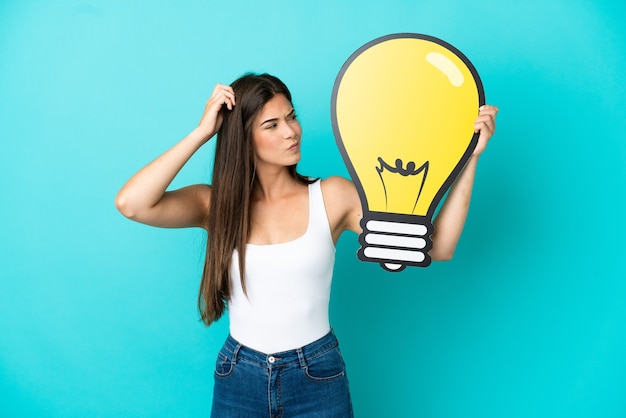 Young Brazilian woman isolated on blue background holding a bulb icon and having doubts