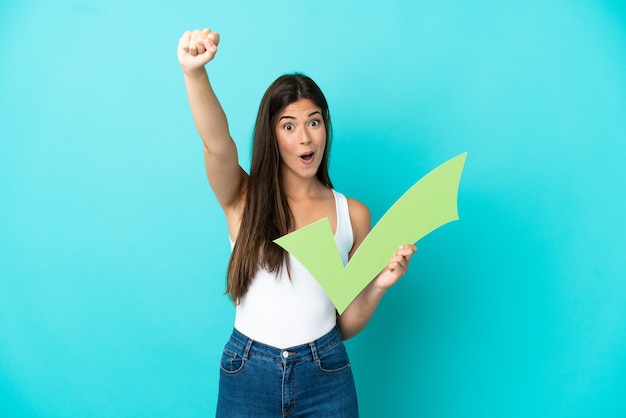 Young Brazilian woman isolated on blue background holding a check icon and celebrating a victory