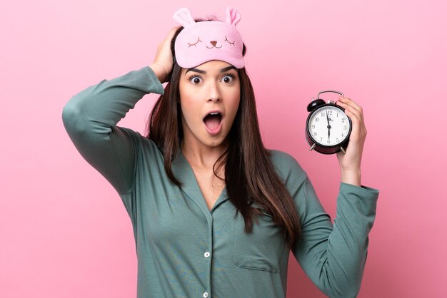 Young Brazilian woman isolated on pink background in pajamas and holding clock with surprised expression