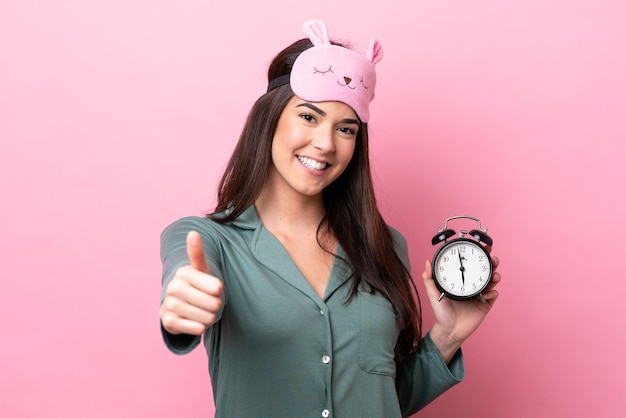 Young Brazilian woman isolated on pink background in pajamas and holding clock with thumb up