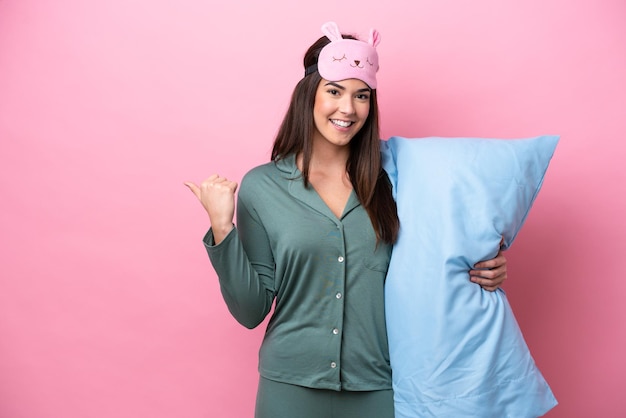 Young Brazilian woman isolated on pink background in pajamas and pointing to the side to present a product