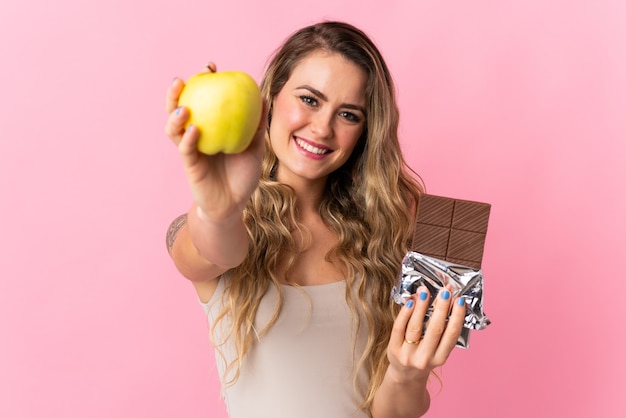 Молодая бразильская женщина изолирована на розовом, принимая шоколадную таблетку в одной руке и яблоко в другой