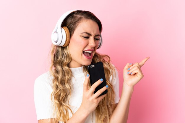 Young brazilian woman isolated on pink wall listening music with a mobile and singing