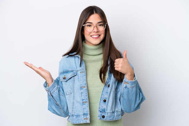 Young Brazilian woman isolated on white background holding copyspace imaginary on the palm to insert an ad and with thumbs up