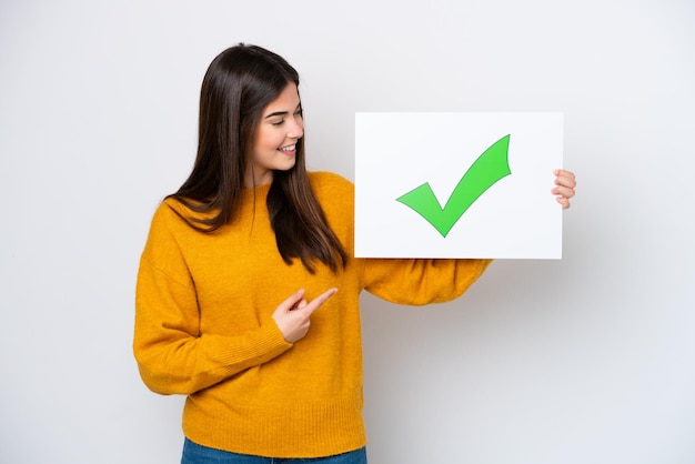 Young Brazilian woman isolated on white background holding a placard with text Green check mark icon and pointing it