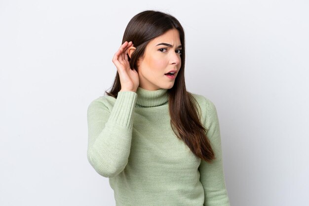 young brazilian woman isolated on white background listening to something by putting hand on the ear