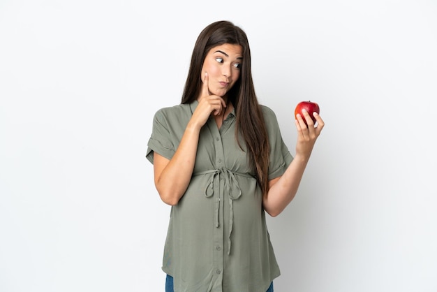 Young Brazilian woman isolated on white background pregnant and holding an apple while thinking