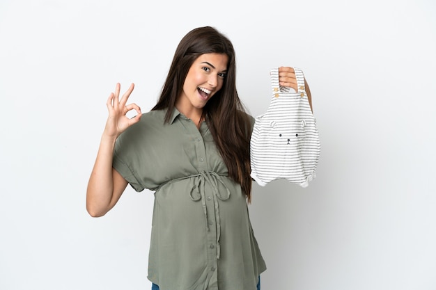 Young Brazilian woman isolated on white background pregnant and holding baby clothes with ok sign