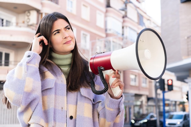 Young Brazilian woman at outdoors holding a megaphone and having doubts