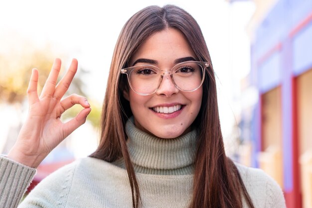 Молодая бразильянка на улице в очках и делает знак ОК