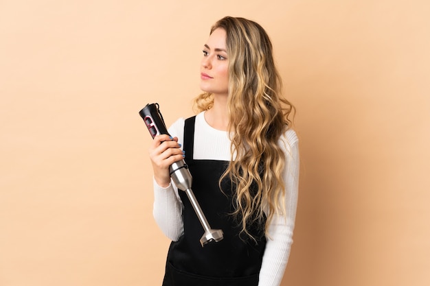 Young brazilian woman using hand blender isolated on beige looking to the side