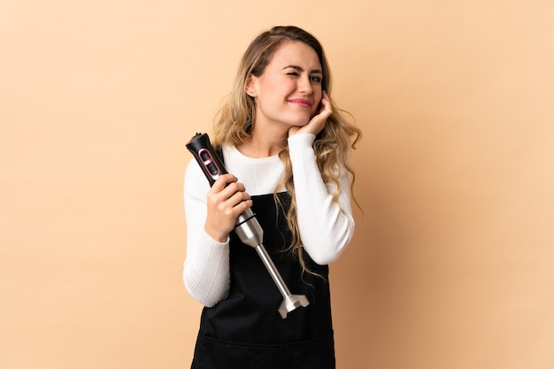 Young brazilian woman using hand blender isolated on beige wall frustrated and covering ears