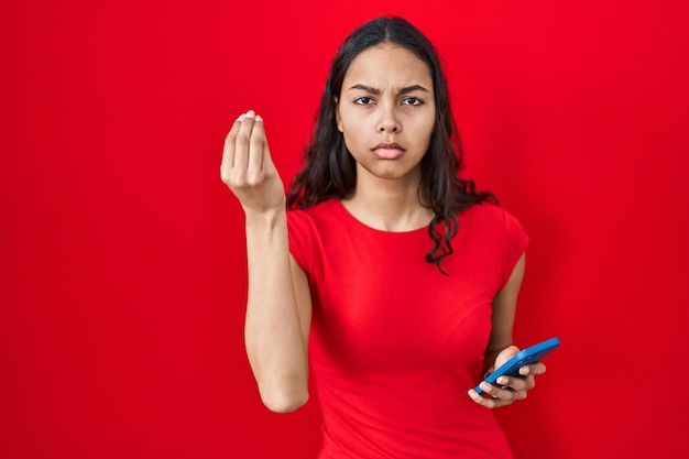 赤い背景の上にスマートフォンを使用して、手と指の自信に満ちた表情でイタリアのジェスチャーをしている若いブラジル人女性