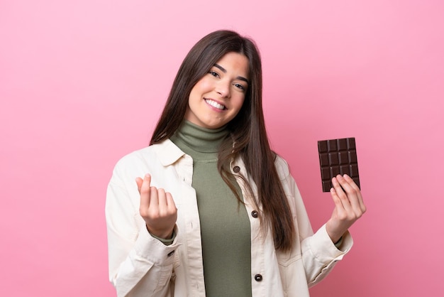 Young Brazilian woman with chocolat isolated on pink background making money gesture