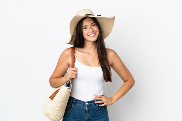 Giovane donna brasiliana con pamela in possesso di una borsa da spiaggia isolata su sfondo bianco in posa con le braccia all'anca e sorridente