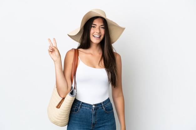 Young Brazilian woman with Pamela holding a beach bag isolated on white background smiling and showing victory sign