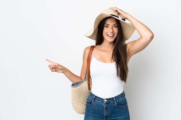 Giovane donna brasiliana con pamela che tiene in mano una borsa da spiaggia isolata su sfondo bianco sorpresa e che punta il dito di lato