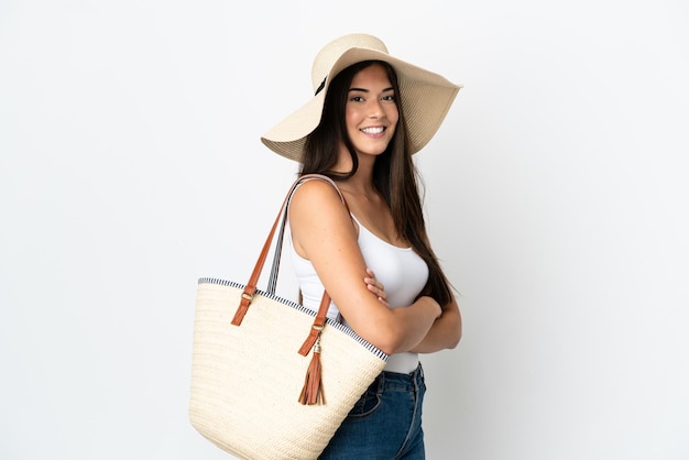 Giovane donna brasiliana con pamela che tiene in mano una borsa da spiaggia isolata su sfondo bianco con le braccia incrociate e guardando avanti