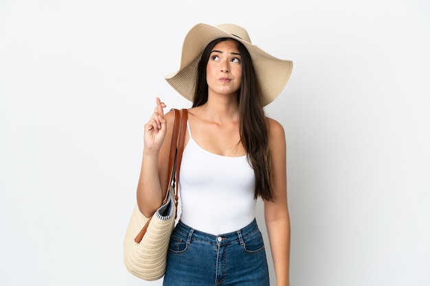 Young Brazilian woman with Pamela holding a beach bag isolated on white background with fingers crossing and wishing the best