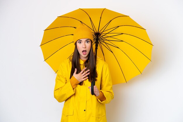 防雨コートと白い背景で隔離の傘を持つ若いブラジル人女性は、右を見ながら驚いてショックを受けました