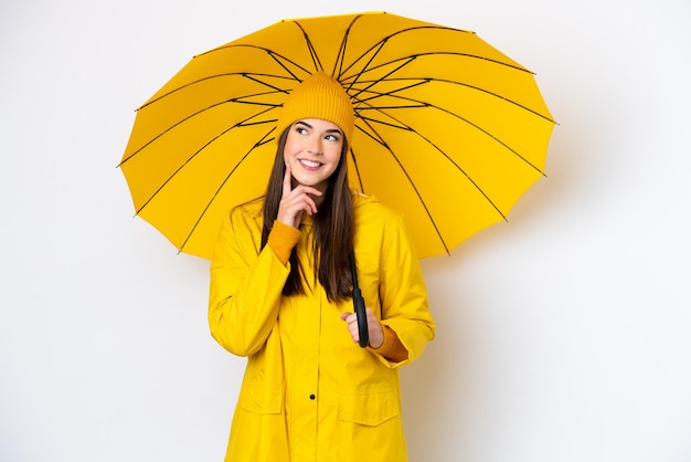 Young Brazilian woman with rainproof coat and umbrella isolated on white background thinking an idea while looking up