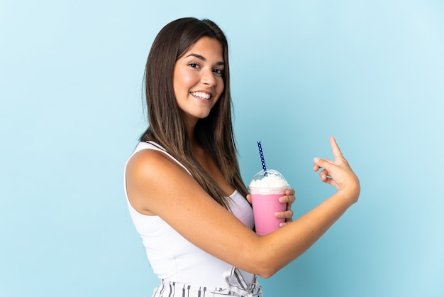 Young brazilian woman with strawberry milkshake isolated on blue wall pointing back