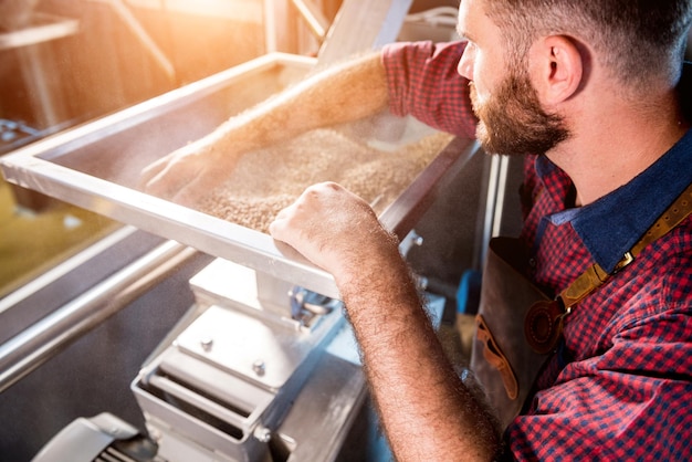 A young brewer in a leather apron controls the grinding of malt seeds in a mill at a modern brewery