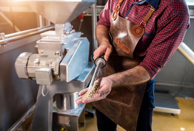 Un giovane birraio in un grembiule in pelle controlla la macinazione dei semi di malto in un mulino in una moderna fabbrica di birra