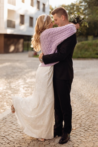 Young bride in bridal dress lilac cardigan with bandeau on head stand holding bouquet hugging brideg