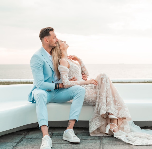 Young bride fashionable blonde model in luxury wedding dress\
with groom on the coast couple walks at the pier on sunrise