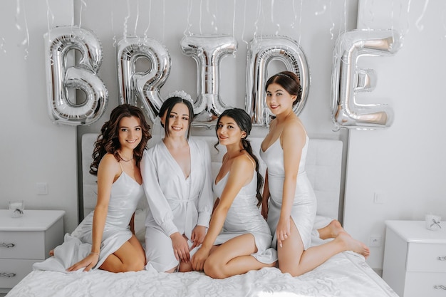 Foto giovani damigelle di sposa in abiti bianchi di seta su un letto nella stanza delle spose belle donne che festeggiano la festa di sposa sedute sul letto