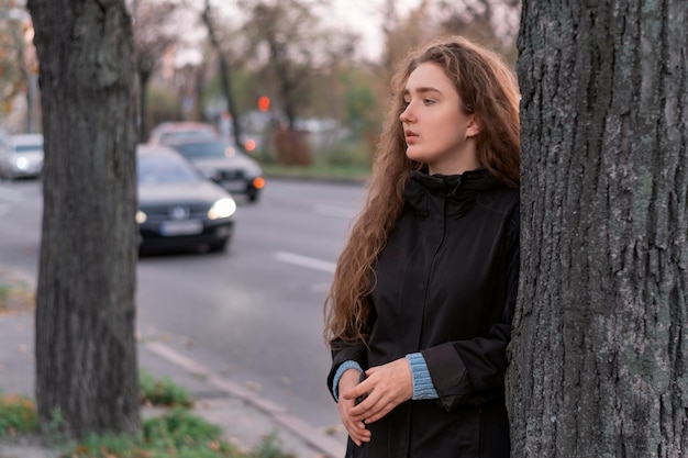 曇りの秋の日に高速道路の近くの木のそばに立っている若い陰気な女性道路の背景にコートを着た十代の少女