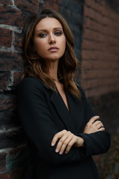 Young brown hair styling fashion woman with beautiful face in black classic coat with opened neck standing in pose with crossed on chest hands arms on the city street with red brick fabric loft wall