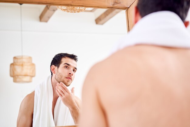 Молодой брюнет с белым полотенцем на плече стоит возле зеркала в ванне утром. Вернуться крупным планом