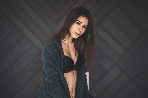 Young brunette in black underwear, knitted knee pads and cardigan sits on a wooden chair.