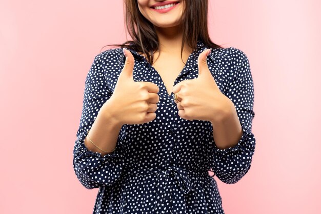 Young brunette in a blue dress with white polka dots showing thumbs up closeup and smiling at camera on pink background
