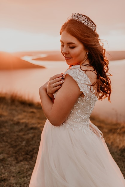 Young brunette bride in white wedding dress with a crown on her head stands on cliff