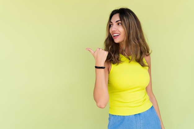 Young brunette in casual wear isolated on green background happy look to camera smile excited point