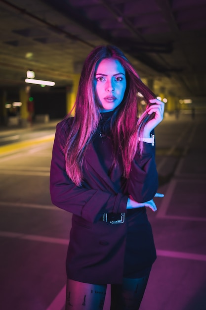 Young brunette Caucasian girl at night in an underground parking lot, illuminated with neon lights