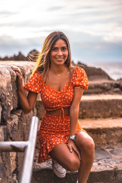 Zumaia, Gipuzkoa 마을 Itzurrun 해변에 빨간 드레스에서 젊은 갈색 머리 백인 여자. 바스크 지방. 계단에서 바다를 즐기는 라이프 스타일 세션