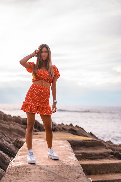 Photo a young brunette caucasian woman in a red dress on the beach of itzurrun in the town of zumaia, gipuzkoa. basque country. lifestyle session, perched on a walkway by the sea