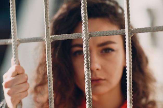 Young brunette curly woman in orange suit behind jail bars Female in colorful overalls portrait