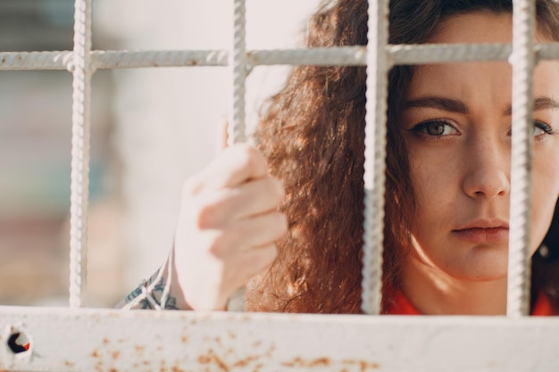 Young brunette curly woman in orange suit behind jail bars Female in colorful overalls portrait