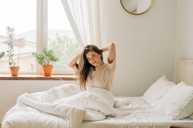 A young brunette enjoying awakening sits on a bed on bed linen near a window in a cozy apartment