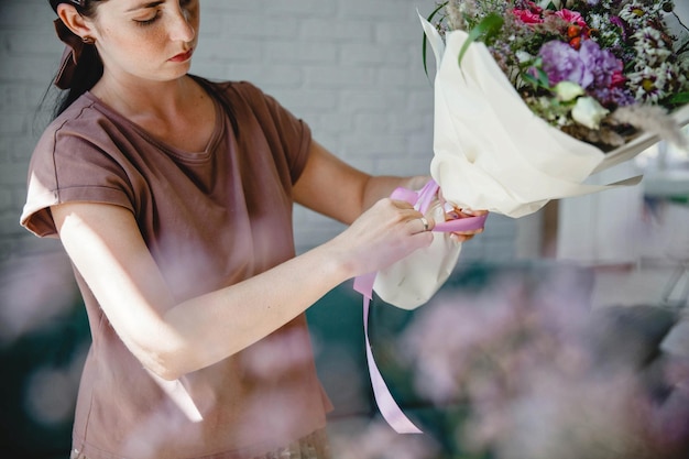 若いブルネットのヨーロッパ人女性の花屋は、彼女のワーク ショップでリボンで紙に包まれたブーケを結ぶ