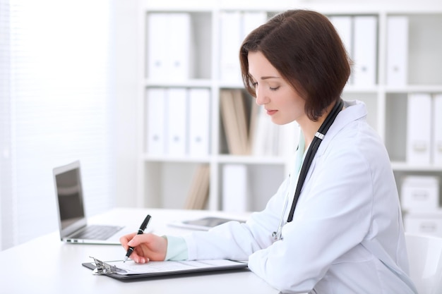 Young brunette female doctor is  sitting at the table and  filling up medical application form.  Health care, insurance and help concept.