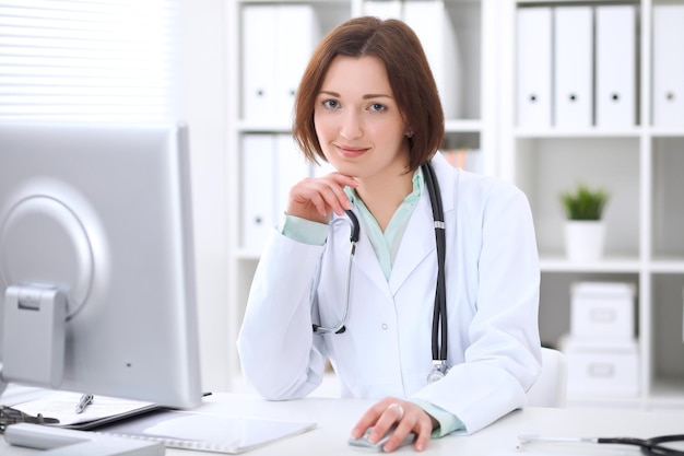 Young brunette female doctor sitting at a desk and working on the computer at the hospital office.  Health care, insurance and help concept. Physician ready to examine patient