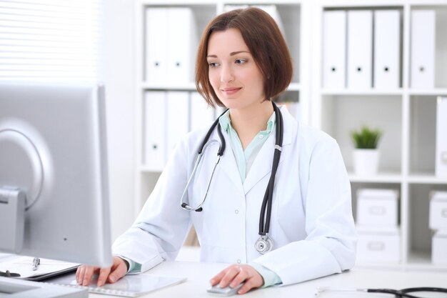 Young brunette female doctor sitting at a desk and working on the computer at the hospital office