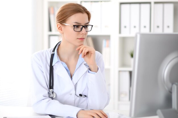 Young brunette female doctor sitting at the table and working by computer  at hospital office.    Doctor is in trouble with diagnosis. Health care, insurance and help concept.