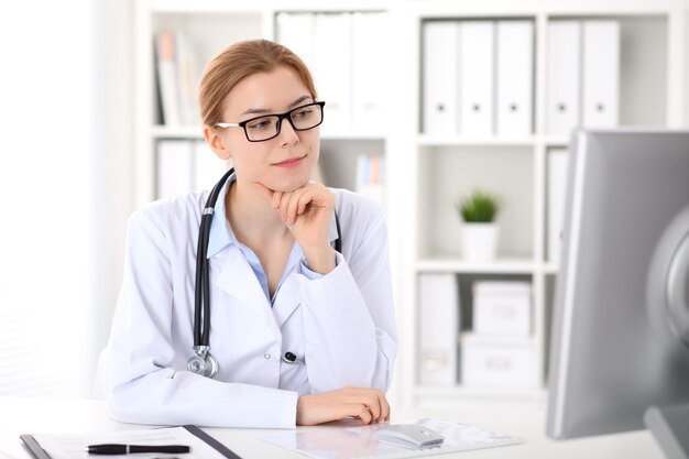 Young brunette female doctor sitting at the table and working by computer  at hospital office.    Doctor is in trouble with diagnosis. Health care, insurance and help concept.